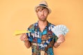 Young blond man wearing summer style holding paper plane and dollars puffing cheeks with funny face Royalty Free Stock Photo