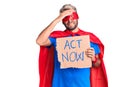 Young blond man wearing super hero costume holding act now cardboard banner stressed and frustrated with hand on head, surprised