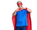 Young blond man wearing super hero custome smiling in love doing heart symbol shape with hands