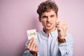 Young blond romantic man with curly hair holdiing paper with love message for valentines day annoyed and frustrated shouting with