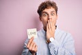 Young blond romantic man with curly hair holdiing paper with love message for valentines day cover mouth with hand shocked with