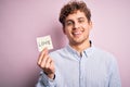 Young blond romantic man with curly hair holdiing paper with love message for valentines day with a happy face standing and