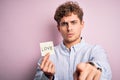 Young blond romantic man with curly hair holdiing paper with love message for valentines day pointing with finger to the camera