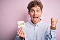 Young blond romantic man with curly hair holdiing paper with love message for valentines day screaming proud and celebrating