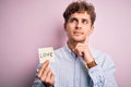 Young blond romantic man with curly hair holdiing paper with love message for valentines day serious face thinking about question,