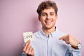 Young blond romantic man with curly hair holdiing paper with love message for valentines day with surprise face pointing finger to