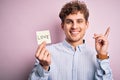 Young blond romantic man with curly hair holdiing paper with love message for valentines day surprised with an idea or question