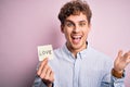 Young blond romantic man with curly hair holdiing paper with love message for valentines day very happy and excited, winner