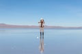 Young blond travel girl with thumbs up stay in water mirror with blue sky reflection in salt flat Uyuni