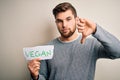 Young blond veggie man with beard and blue eyes holding paper with vegan message with angry face, negative sign showing dislike