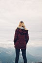 Young blond woman facing away standing on a cliff in winter wearing dark red burgundy coat looking in the distance to mountains