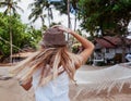 Young blond woman fluttering in hat standing with her back on tropical resort with hammock enjoying vacation