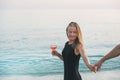 Young blond woman with glass of rose wine holding man's hand on beach by the sea at sunset. Alanya, Turkey.