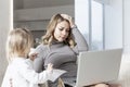 Young blond woman with laptop on the sofa with her baby. Remote work stress