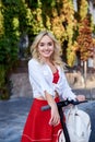 Young blond woman, posing with black electric scooter in city center old historical building covered with green plants. Summer Royalty Free Stock Photo