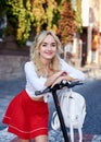 Young blond woman, posing with black electric scooter in city center old historical building covered with green plants. Summer Royalty Free Stock Photo