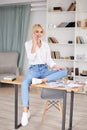 Young blond woman, wearing casual clothes, sitting on table, talking on mobile phone. Freelancer employed at home. Work process in Royalty Free Stock Photo