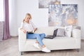 Young blond woman, wearing casual jeans and white top, sitting on sofa, working on design project on laptop. Freelancer employed Royalty Free Stock Photo