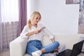 Young blond woman, wearing casual jeans and white top, sitting on sofa, working on design project on laptop. Freelancer employed Royalty Free Stock Photo