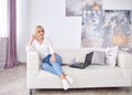 Young blond woman, wearing casual jeans and white top, sitting on sofa, working on design project on laptop. Freelancer employed Royalty Free Stock Photo