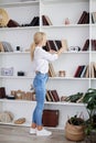 Young blond woman, wearing casual jeans and white top, standing near book shelves in the office room. Freelancer employed at home Royalty Free Stock Photo