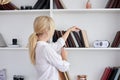 Young blond woman, wearing casual jeans and white top, standing near book shelves in the office room. Freelancer employed at home Royalty Free Stock Photo