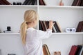 Young blond woman, wearing casual jeans and white top, standing near book shelves in the office room. Freelancer employed at home Royalty Free Stock Photo