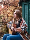 Young blond woman, wearing colorful cardigan, holding acoustic guitar, sitting on a bench in front of old green wooden hut in the Royalty Free Stock Photo