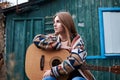 Young blond woman, wearing colorful cardigan, holding acoustic guitar, sitting on a bench in front of old green wooden hut in the Royalty Free Stock Photo