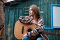 Young blond woman, wearing colorful cardigan, holding acoustic guitar, sitting on a bench in front of old green wooden hut in the Royalty Free Stock Photo