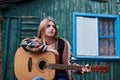 Young blond woman, wearing colorful cardigan, holding acoustic guitar, sitting on a bench in front of old green wooden hut in the Royalty Free Stock Photo