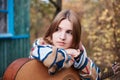 Young blond woman, wearing colorful cardigan, leaning on acoustic guitar, thinking,sitting in front of old green wooden hut in the Royalty Free Stock Photo