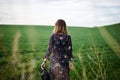 Young blond woman, wearing long dark boho dress, holding black hat standing with back to camera in green field in spring. Model Royalty Free Stock Photo