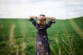 Young blond woman, wearing long dark boho dress, holding black hat standing with back to camera in green field in spring. Model Royalty Free Stock Photo