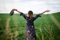 Young blond woman, wearing long dark boho dress, holding black hat standing with back to camera in green field in spring. Model Royalty Free Stock Photo