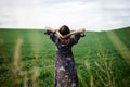 Young blond woman, wearing long dark boho dress, holding black hat standing with back to camera in green field in spring. Model Royalty Free Stock Photo