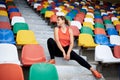 Young blond woman, wearing orange top, black leggings, orange sneakers, sitting on the stairs on stadium tribune, relaxing after