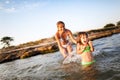 Young blond woman in white bikini and small happy girl having fun in sea water with rocky beach at background Royalty Free Stock Photo