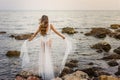Young blond woman in white summer dress standing on the rocks and looking at the sea. Caucasian girl enjoys beautiful view at sunr Royalty Free Stock Photo