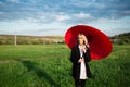 Young blonde beautiful pretty girl, holding red umbrella, wearing black and white suit. Background of cloudy sky and green field. Royalty Free Stock Photo