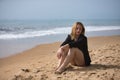 Young, blonde, beautiful woman in a bikini and black colored shirt sitting on the beach, sad, lonely, sorry, depressed. Concept Royalty Free Stock Photo