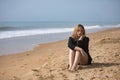 Young, blonde, beautiful woman in a bikini and black colored shirt sitting on the beach, sad, lonely, sorry, depressed. Concept Royalty Free Stock Photo