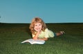 A young blonde boy is lying on the green grass outdoors reading a white book in summer. Smart child reading book, laying Royalty Free Stock Photo