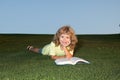 A young blonde boy is lying on the green grass outdoors reading a white book in summer. Smart child reading book, laying Royalty Free Stock Photo