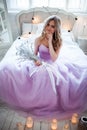 A young blonde bride in a magnificent purple dress sitting on the bed. In the room candles. Girl holds in the hands of