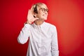 Young blonde business woman with short hair wearing glasses over red background smiling with hand over ear listening an hearing to Royalty Free Stock Photo