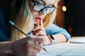 Young blonde caucasian student in eyeglasses thinking and writing something with pencil Royalty Free Stock Photo