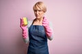 Young blonde cleaner woman with short hair wearing apron and gloves holding scrub scourer annoyed and frustrated shouting with