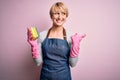 Young blonde cleaner woman with short hair wearing apron and gloves holding scrub scourer pointing and showing with thumb up to