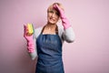 Young blonde cleaner woman with short hair wearing apron and gloves holding scrub scourer stressed with hand on head, shocked with